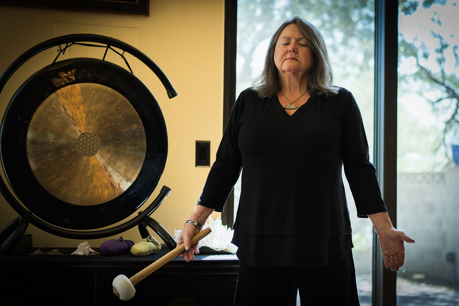 CJ Walker standing in the temple room beside the gong, eyes closed, a mallet in one hand and the other palm open